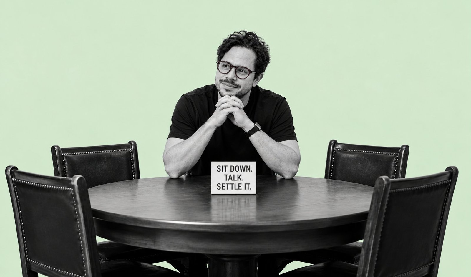 Michael Philippou seated at a round table with four empty leather chairs and a sign reading Sit down. Talk. Settle it.